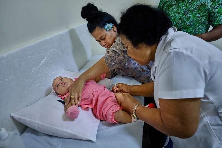 A baby is vaccinated by a nurse while her mother holds the baby's arms.